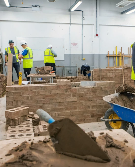 Students in high-visibility vests working in a spacious brickwork workshop, with one student loading mortar into a wheelbarrow as others collaborate on projects. Students in high-visibility vests working in a spacious brickwork workshop, with one student loading mortar into a wheelbarrow as others collaborate on projects.